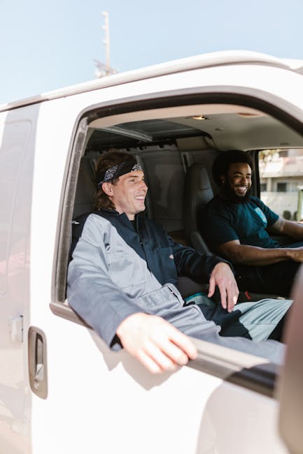 A close-up view from outside a white delivery van shows two men inside, both smiling and engaging in conversation. The man on the left has medium-length hair tied back with a bandana, wearing a light